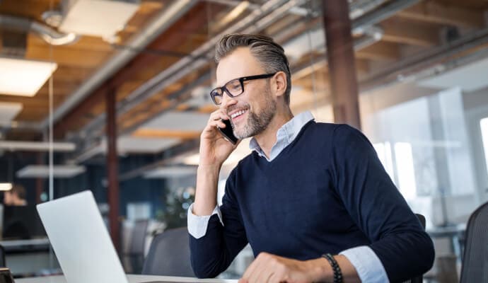 Smiling man with mobile phone held to his ear as he looks at a laptop